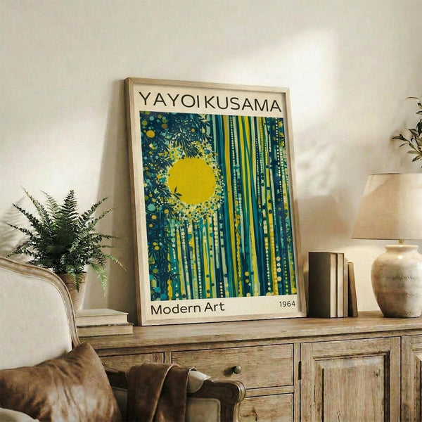 May include: Framed art print with yellow sun and vertical blue-green streaks in polka dot pattern, displayed on rustic wooden sideboard beside fern plant, books, and ceramic vases in bright living room setting with beige sofa and white walls