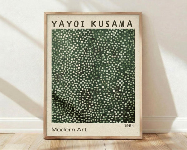 May include: Framed poster print with light wood frame leaning against white wall on wooden floor. Features green background covered in white polka dots of varying sizes creating optical illusion pattern. Text reads "YAYOI KUSAMA" at top, "1984" and "Modern Art" at bottom on cream colored border.