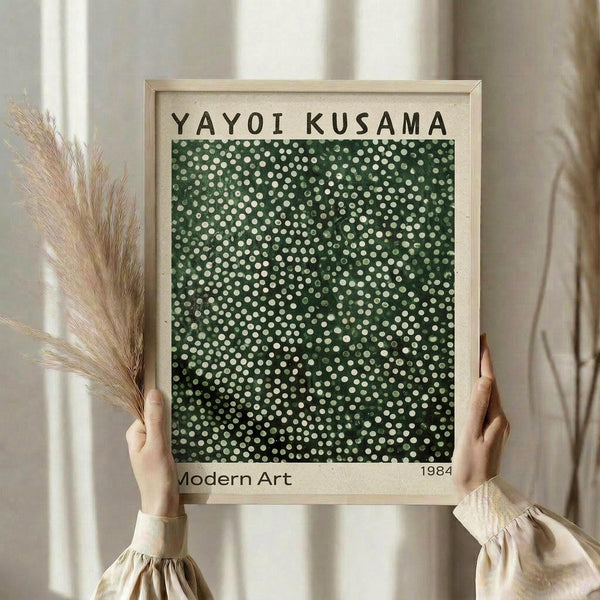 May include: Person holding framed art print featuring green background with scattered white polka dots in varying sizes. Light wood frame with cream matting displays "YAYOI KUSAMA" at top and "Modern Art 1984" at bottom. Hands wear beige ruffled sleeves, dried pampas grass visible on left side against soft neutral curtained background.