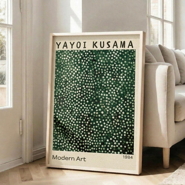 May include: Framed art print with green background covered in white dots of varying sizes, text reading "YAYOI KUSAMA" at top and "Modern Art 1984" at bottom, displayed on light wood floor against white wall next to beige upholstered chair in bright room with large windows