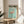 May include: Framed Japanese art exhibition poster featuring pink flamingo on turquoise background, displayed on round wooden side table next to beige upholstered chair. Natural lighting creates soft shadows on white wall. Green plant leaves visible in corner. Poster shows vintage-style typography and exhibition dates from 2007.