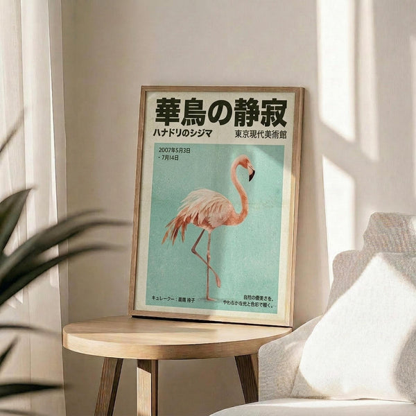 May include: Framed Japanese art exhibition poster featuring pink flamingo on turquoise background, displayed on round wooden side table next to beige upholstered chair. Natural lighting creates soft shadows on white wall. Green plant leaves visible in corner. Poster shows vintage-style typography and exhibition dates from 2007.