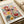 May include: A framed botanical art print featuring stylized flowers, leaves, and plants in vibrant colors including red, yellow, teal, pink, and orange. The artwork displays various floral motifs with dotted patterns and radiating lines in a mid-century modern illustration style. Text reads "YAYOI KUSAMA" at top, "1966" and "Modern Art" at bottom. The print is shown in a light wooden frame resting against neutral-toned paper or fabric backdrop.