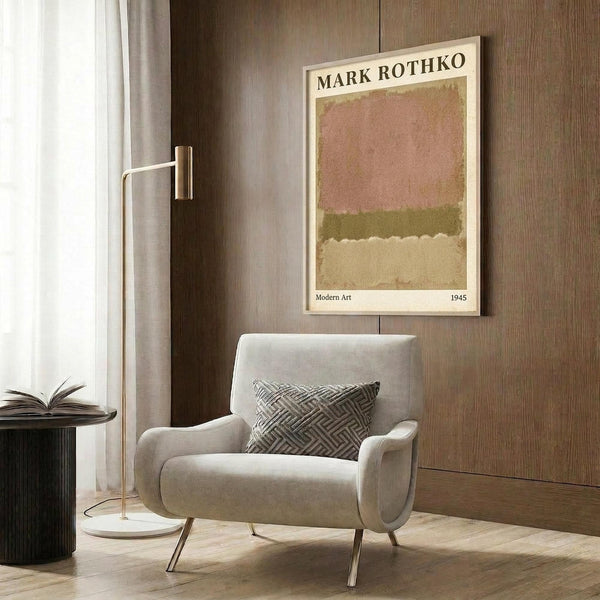 May include: A framed reproduction poster of a Mark Rothko abstract painting featuring horizontal bands of muted pink, olive green, and cream colors. The poster hangs on a wood-paneled wall above a beige upholstered armchair with geometric patterned pillow. A brass floor lamp and small side table with open book complete the modern living room setting with warm neutral tones throughout.