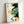 May include: Framed abstract art print leaning against white wall, featuring flowing organic shapes in emerald green, golden yellow, and navy blue with Japanese calligraphy text on left side. Light wood frame with white matting, positioned on light surface with natural lighting casting soft shadows.