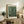 May include: Framed art print featuring green background with white polka dots in varying sizes, displayed on wooden console table alongside potted fern, ceramic vases, and stacked books. Light wooden frame matches rustic furniture. Warm, neutral-toned room setting with beige armchair visible in corner.