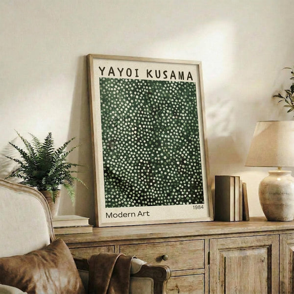 May include: Framed art print featuring green background with white polka dots in varying sizes, displayed on wooden console table alongside potted fern, ceramic vases, and stacked books. Light wooden frame matches rustic furniture. Warm, neutral-toned room setting with beige armchair visible in corner.