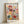 May include: Framed art print featuring colorful botanical illustration with stylized flowers and plants in red, yellow, teal, pink, and purple tones against cream background. The design shows whimsical blooms with dotted patterns, radiating lines, and organic shapes in mid-century modern style. Light wood frame displayed leaning against white wall, showing natural lighting and shadows.