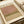 May include: Abstract art print with soft pink and olive green horizontal color fields in minimalist style, displayed in an open art book or catalog. The artwork shows muted earth tones with organic, cloud-like edges between color blocks. White ribbon bookmark visible alongside cream-colored pages in warm, natural lighting.