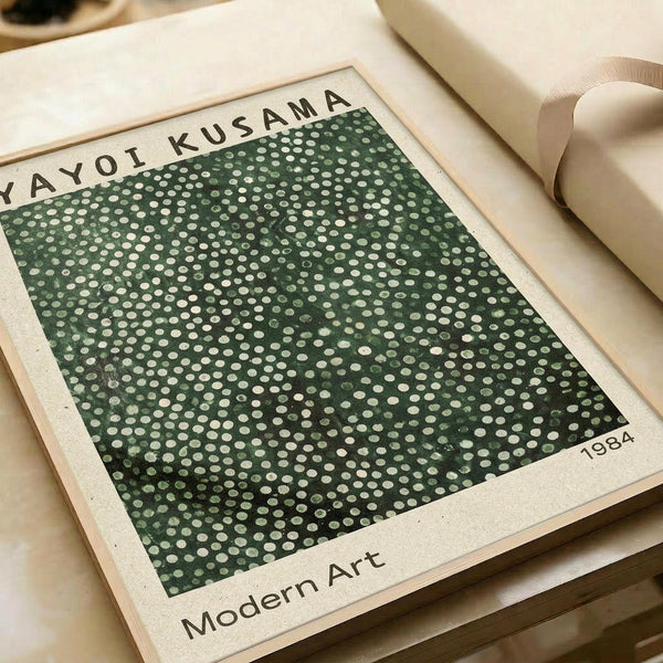 Vintage-style art print featuring Yayoi Kusama's signature polka dot pattern in dark green with cream-colored dots scattered across the surface. The print has "YAYOI KUSAMA" at the top, "1984" in the bottom right corner, and "Modern Art" at the bottom. The poster is displayed on cream-colored paper with a beige ribbon visible in the corner, creating a minimalist aesthetic against neutral packaging materials.