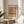 May include: Framed abstract art print in warm earth tones - browns, pinks, greens, and creams in soft rectangular color blocks. Print sits on round wooden side table next to beige upholstered chair with white pillow. Natural sunlight streams across white walls, with plant leaves visible in corner. Thin wood frame surrounds poster with white matting.