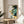 May include: A framed abstract art print with flowing organic shapes in navy blue, teal, gold, and cream colors alongside Japanese calligraphy text, displayed on a light wood round side table next to a beige upholstered chair. Natural sunlight creates soft shadows on the white wall behind. A plant's leaves are partially visible in the corner, creating an elegant minimalist home decor scene.