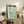 May include: Framed Japanese exhibition poster featuring pink flamingo on turquoise background, displayed on rustic wooden sideboard in living room setting with fern plant, ceramic vases, books, and brown leather armchair visible in warm neutral toned interior space.