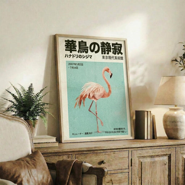 May include: Framed Japanese exhibition poster featuring pink flamingo on turquoise background, displayed on rustic wooden sideboard in living room setting with fern plant, ceramic vases, books, and brown leather armchair visible in warm neutral toned interior space.