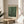 May include: Framed art print with green background covered in white polka dots, displayed on round wooden side table next to beige upholstered chair. Natural lighting creates soft shadows on white wall. Plant leaves visible in corner. Poster shows "YAYOI KUSAMA" at top, "Modern Art" and "1984" at bottom in simple black text on cream-colored paper with natural wood frame.