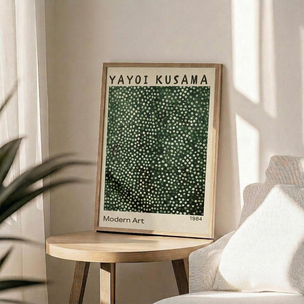 May include: Framed art print with green background covered in white polka dots, displayed on round wooden side table next to beige upholstered chair. Natural lighting creates soft shadows on white wall. Plant leaves visible in corner. Poster shows "YAYOI KUSAMA" at top, "Modern Art" and "1984" at bottom in simple black text on cream-colored paper with natural wood frame.