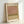 May include: Framed abstract art print leaning against white wall corner. The artwork features soft horizontal bands of muted colors - peachy pink at top transitioning to olive green in middle and sandy beige at bottom with white cloudy area between layers. Light wooden frame surrounds cream-colored matting with "MARK ROTHKO" text at top, "1945" and "Modern Art" at bottom. Soft natural lighting creates gentle shadows.