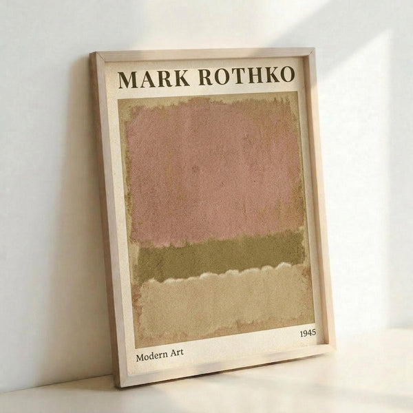 May include: Framed abstract art print leaning against white wall corner. The artwork features soft horizontal bands of muted colors - peachy pink at top transitioning to olive green in middle and sandy beige at bottom with white cloudy area between layers. Light wooden frame surrounds cream-colored matting with "MARK ROTHKO" text at top, "1945" and "Modern Art" at bottom. Soft natural lighting creates gentle shadows.