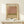 May include: Framed art print in light wood frame leaning against white wall on wooden shelf. Print features horizontal bands of muted pink, olive green, and beige colors in abstract expressionist style. Small white gift box with evergreen sprig sits in front of the frame. Soft natural lighting creates gentle shadows on the minimal, neutral-toned display.