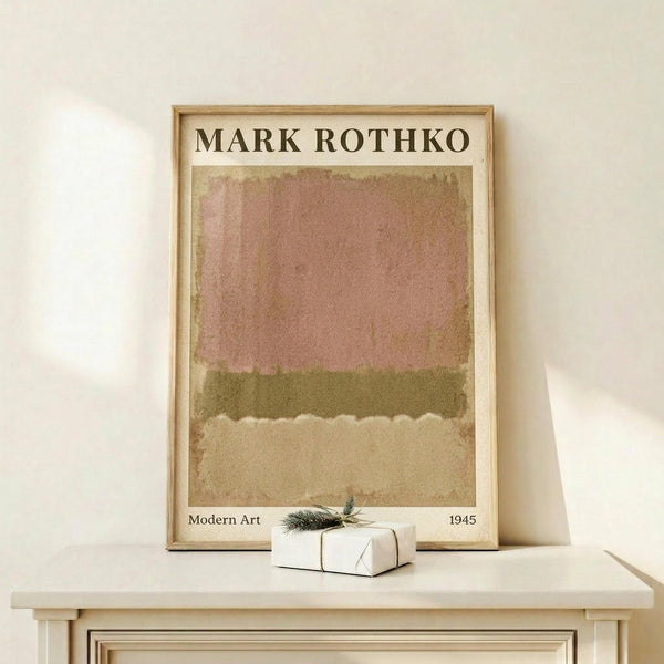 May include: Framed art print in light wood frame leaning against white wall on wooden shelf. Print features horizontal bands of muted pink, olive green, and beige colors in abstract expressionist style. Small white gift box with evergreen sprig sits in front of the frame. Soft natural lighting creates gentle shadows on the minimal, neutral-toned display.