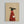 Minimalist illustration of a black greyhound wearing a red ribbed sweater, holding a white coffee mug with steam rising above. The dog is shown in profile against a tan textured background with "Brewed for Good Dogs" text at the top. The artwork is displayed in a light wood frame hanging on a white wall.