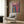 May include: Modern reading corner with beige upholstered armchair featuring geometric throw pillow, brass floor lamp, dark round side table with open book, and colorful abstract poster on wood-paneled wall. The vibrant artwork displays swirling rainbow patterns with an orange figure silhouette against flowing multicolored waves and Japanese characters at the top.
