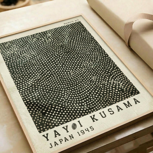 A minimalist art print featuring a black square filled with thousands of small white polka dots in varying densities, creating an optical illusion effect. The dots appear denser toward the center and gradually become more sparse toward the edges. Text reads "modern art" at the top and "YAYOI KUSAMA JAPAN 1945" at the bottom in black letters on cream paper. The print is displayed flat on a light wooden surface next to rolled cream-colored paper tubes.