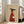 May include: Framed poster print leaning against white wall next to beige upholstered chair in bright living room with large windows. Artwork shows stylized black greyhound wearing red ribbed sweater, holding white coffee cup with steam rising. Text reads "Brewed for Good Dogs" on tan textured background. Light wood frame, approximately 2-3 feet tall, casting soft shadow on white wooden floor.