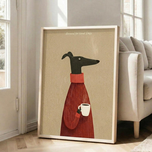 May include: Framed poster print leaning against white wall next to beige upholstered chair in bright living room with large windows. Artwork shows stylized black greyhound wearing red ribbed sweater, holding white coffee cup with steam rising. Text reads "Brewed for Good Dogs" on tan textured background. Light wood frame, approximately 2-3 feet tall, casting soft shadow on white wooden floor.
