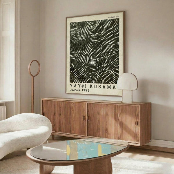 May include: Black and white polka dot art print in wooden frame hanging above mid-century walnut credenza in minimalist living room. Modern white curved chair and oval glass coffee table with blue and yellow artwork visible. Cream walls, natural lighting, contemporary Scandinavian-style interior design with warm wood tones throughout.