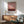 May include: Abstract art print in warm pink and burgundy tones displayed in modern living room setting with mint green armchair, wooden sideboard, ceramic table lamp, and natural sunlight casting shadows on white wall. Print features gradient colors transitioning from light pink sky to darker reddish-brown horizon in minimalist Rothko style, housed in thin wooden frame.