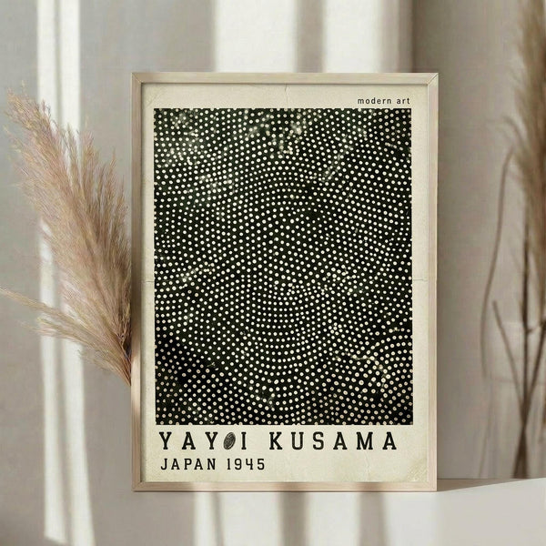May include: Framed poster with black background covered in white dots creating a radial pattern, displayed on light wooden surface next to dried pampas grass stems. The artwork features dense polka dots that appear to emanate from the center, with text below reading "YAYOI KUSAMA JAPAN 1945" in black letters. Light wooden frame against cream curtained background in soft natural lighting.
