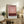 May include: Framed abstract art print with pink and burgundy gradient colors displayed on rustic wooden sideboard. Room scene shows beige upholstered chair with brown leather cushion, potted fern, ceramic vases, and stacked books. Poster features museum-style text layout with atmospheric color blocks transitioning from light pink at top to deep maroon at bottom. Natural lighting creates warm, cozy living room ambiance.