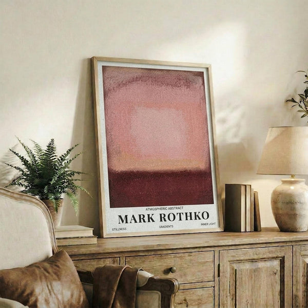 May include: Framed abstract art print with pink and burgundy gradient colors displayed on rustic wooden sideboard. Room scene shows beige upholstered chair with brown leather cushion, potted fern, ceramic vases, and stacked books. Poster features museum-style text layout with atmospheric color blocks transitioning from light pink at top to deep maroon at bottom. Natural lighting creates warm, cozy living room ambiance.