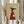 May include: Person in beige clothing holding framed poster showing stylized black greyhound wearing red ribbed sweater with pocket holding white coffee cup, text "Brewed for Good Dogs" at top, tan background, light wood frame, dried pampas grass in left hand, neutral room setting with white curtains