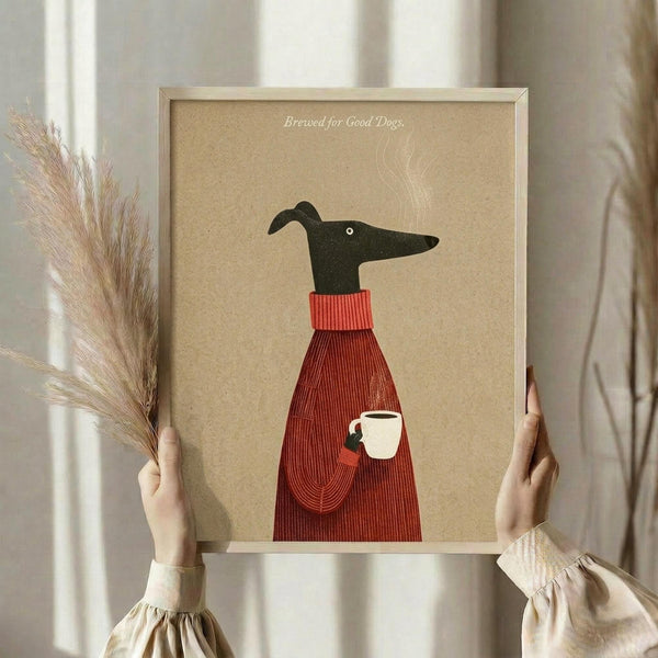 May include: Person in beige clothing holding framed poster showing stylized black greyhound wearing red ribbed sweater with pocket holding white coffee cup, text "Brewed for Good Dogs" at top, tan background, light wood frame, dried pampas grass in left hand, neutral room setting with white curtains