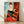 May include: Framed reproduction of Modigliani's portrait painting showing a woman with elongated features, auburn hair in a bun, wearing a black top and red skirt, seated in a red chair against a blue and brown background. The artwork displays characteristic modernist style with simplified forms and warm earth tones, presented in a light wood frame leaning against a white wall on wooden flooring.