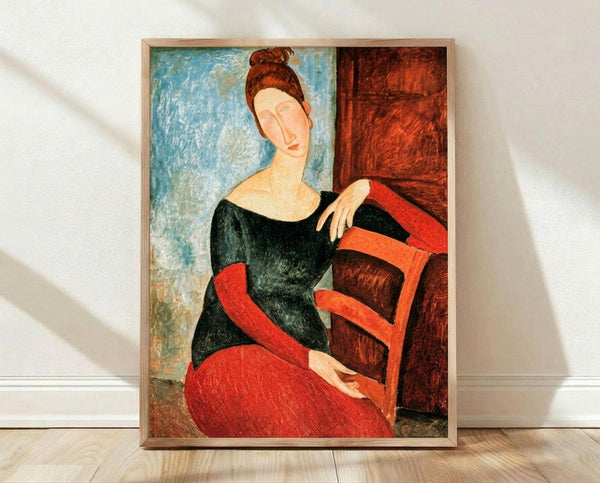 May include: Framed reproduction of Modigliani's portrait painting showing a woman with elongated features, auburn hair in a bun, wearing a black top and red skirt, seated in a red chair against a blue and brown background. The artwork displays characteristic modernist style with simplified forms and warm earth tones, presented in a light wood frame leaning against a white wall on wooden flooring.