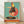 May include: Framed poster print leaning against white wall on light wood floor. Artwork shows stylized rooster with yellow head, red comb, wearing black polka dot dress and sunglasses, standing on vintage turntable. Teal background with large coral circle behind bird. Mid-century modern illustration style with warm retro colors. Natural wood frame, approximately 16x20 size relative to baseboard trim.
