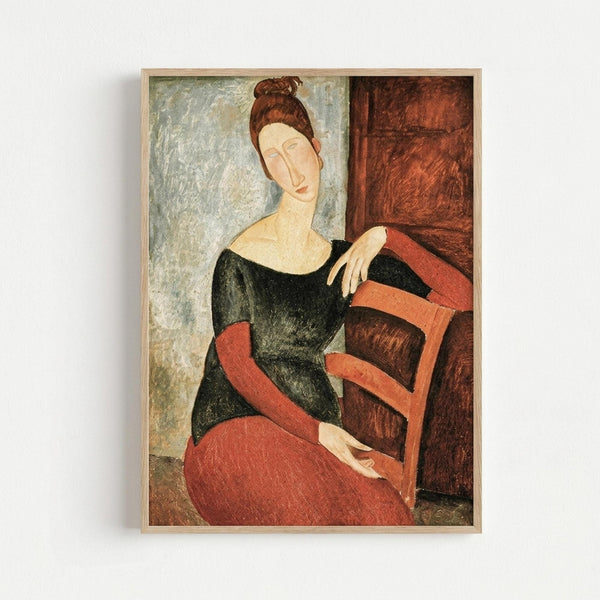 Framed art print showing a woman with auburn hair in a bun, wearing a black top and coral-red skirt, seated in a wooden chair. The painting features elongated proportions and muted earth tones characteristic of early 20th century modernist style. The background shows textured walls in pale green and reddish-brown tones. The artwork is displayed in a simple light wood frame against a white wall.