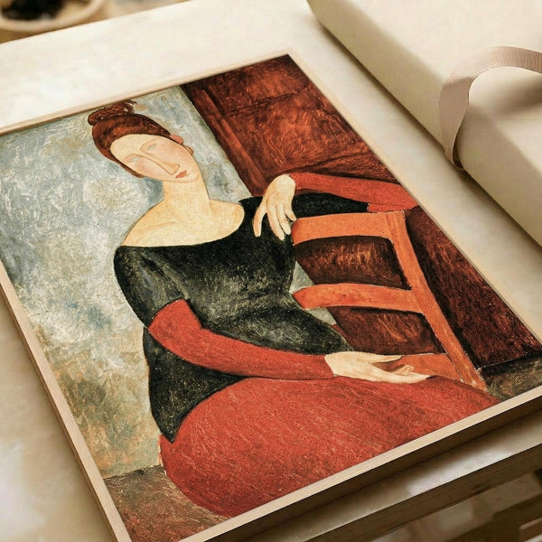 Art print displayed in an open book or portfolio showing a seated woman with reddish-brown hair in an updo, wearing a black top and coral-red skirt. The figure has elongated features characteristic of Modigliani's style, with pale skin and simplified facial features. She sits in a wooden chair against a muted blue-green background. The artwork uses warm earth tones of coral, rust, and deep greens with visible texture suggesting oil paint or pastels.
