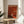 May include: Orange and red minimalist art poster in thin wooden frame, leaning against white wall on round wooden side table next to beige upholstered chair. Natural sunlight creates soft shadows across the scene. Poster features geometric design with small silhouette figure and white text against warm gradient background.