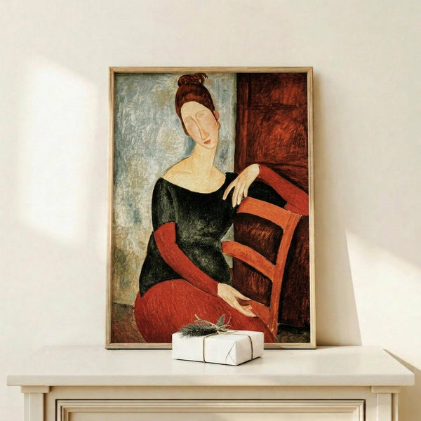 May include: Framed art print showing a woman with auburn hair in a black dress and red skirt, seated in an elongated modernist style. The painting features muted earth tones with blue-gray background. Display shows the artwork on a light wooden surface beside a small white wrapped gift with greenery, all against a cream wall.