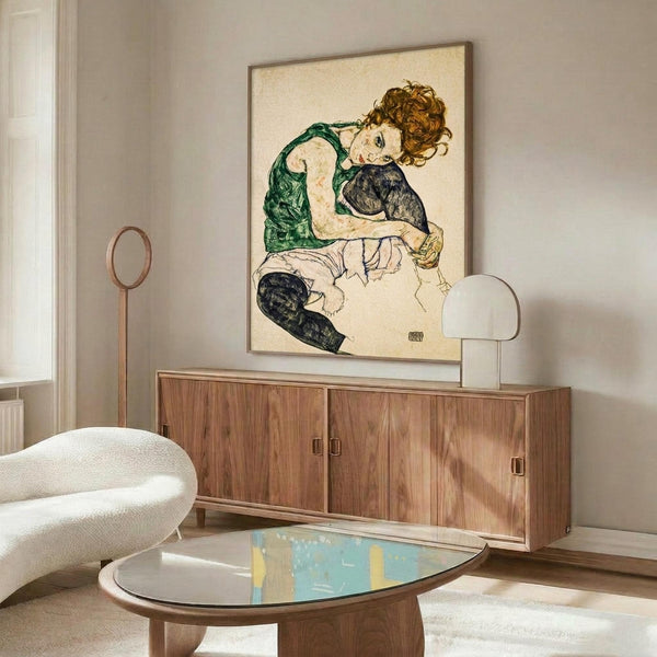 May include: Modern living room interior featuring framed reproduction of early 20th century expressionist artwork showing seated woman with curly auburn hair wearing green sleeveless top and dark stockings against cream background. Wood credenza beneath holds white sculptural lamp, white upholstered chair and glass coffee table with colorful abstract art visible nearby.
