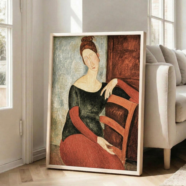 May include: Large framed art print leaning against white wall in bright modern living room with beige sofa and windows. Artwork shows woman with elongated face and brown hair bun, wearing black top and coral red skirt, seated in chair. Painting features Modigliani's distinctive style with muted earth tones and simplified forms. Print has thin light wood frame and appears oversized compared to furniture.