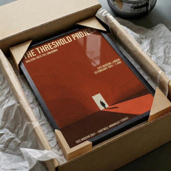May include: Framed poster with minimalist orange-red design featuring silhouetted figure in geometric landscape, packaged in cardboard box with protective tissue paper and wooden frame corners visible, sitting on white surface next to black glass object.