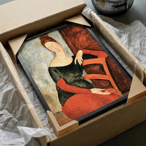 May include: Framed reproduction of Modigliani's portrait showing a woman with elongated neck and stylized features in muted earth tones of rust red, olive green, and cream. The artwork sits in a black frame within an open cardboard shipping box surrounded by white protective tissue paper, with wooden frame corners visible and a glass bowl in the background.