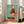 May include: Framed poster print leaning against white wall in bright living room with beige sofa and windows. Artwork shows stylized yellow rooster wearing black polka dot dress with red accents, sunglasses, and red comb, dancing on vinyl record turntable against turquoise background with large coral circle. Retro mid-century modern illustration style with textured appearance in light wood frame.