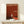 May include: Framed poster with rust-red gradient background featuring silhouette of person walking toward bright doorway opening. White text reads "The Threshold Project" and "Walking into the Unknown" with exhibition details for Tate Modern London. Small wrapped gift with evergreen sprig sits on light wood surface beside frame against cream wall.