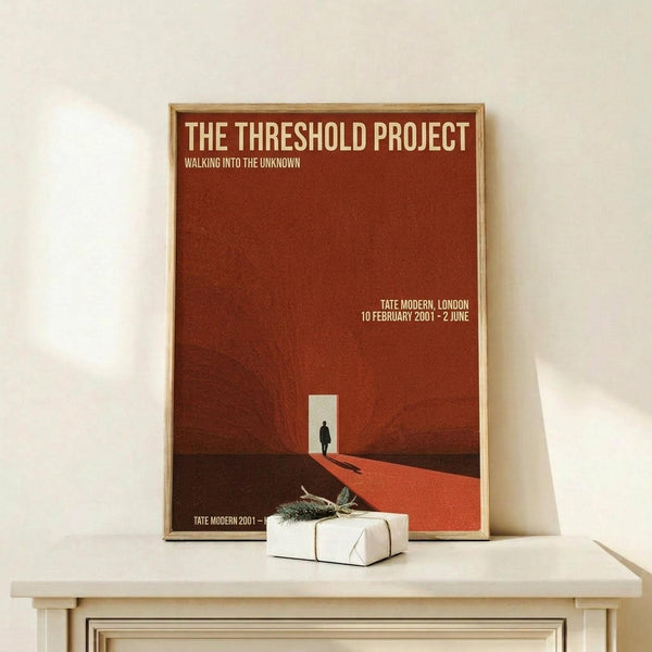May include: Framed poster with rust-red gradient background featuring silhouette of person walking toward bright doorway opening. White text reads "The Threshold Project" and "Walking into the Unknown" with exhibition details for Tate Modern London. Small wrapped gift with evergreen sprig sits on light wood surface beside frame against cream wall.